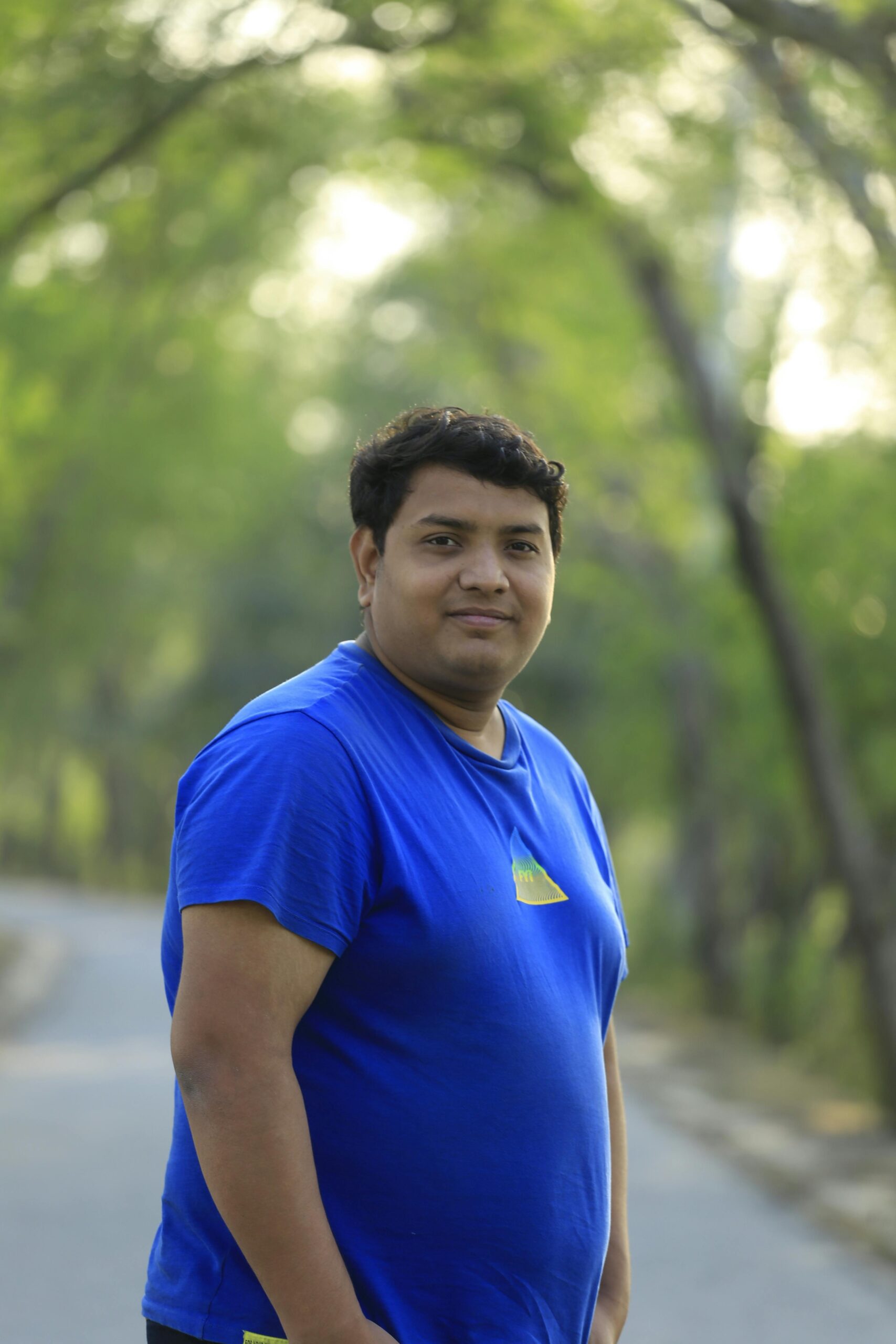 A man in a blue shirt poses on a peaceful park road surrounded by green trees.