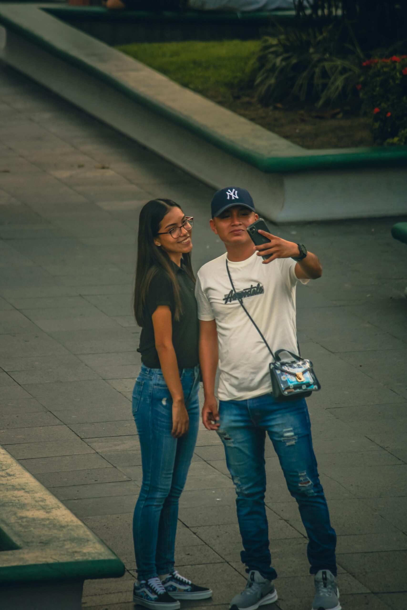 A young couple takes a selfie on an urban walkway, showcasing casual fashion and city life.